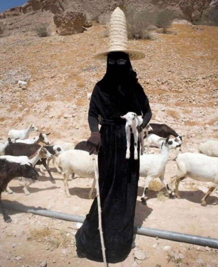 Person wearing tall straw hat and black clothing holding a goat among other goats