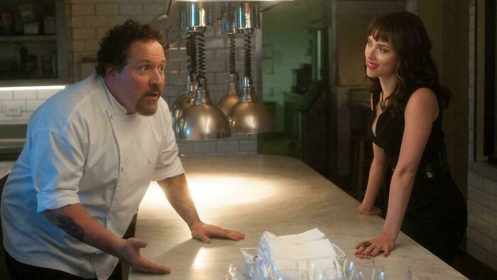 A male chef with a surprised expression talks to a woman in a black dress, both reflecting nonsensical movie details.