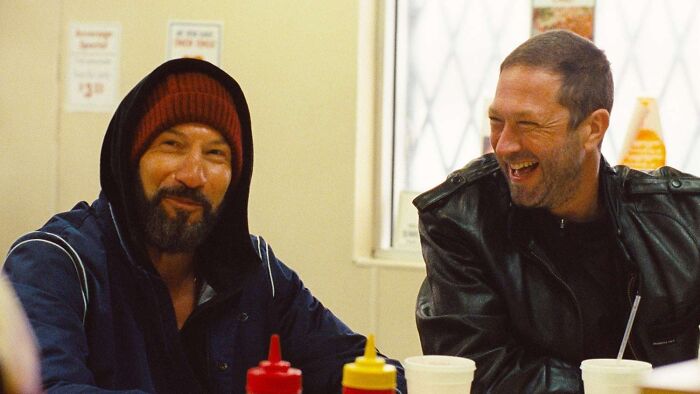Dois homens em um restaurante. Um usa gorro vermelho e moletom azul, o outro uma jaqueta de couro preta, rindo. A antecipação da 5ª temporada do Bear.