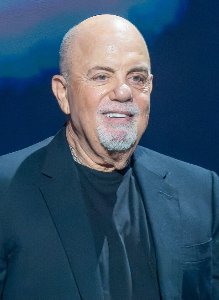 Close-up of Billy Joel smiling, wearing a dark suit jacket and a black shirt, showcasing his iconic career.