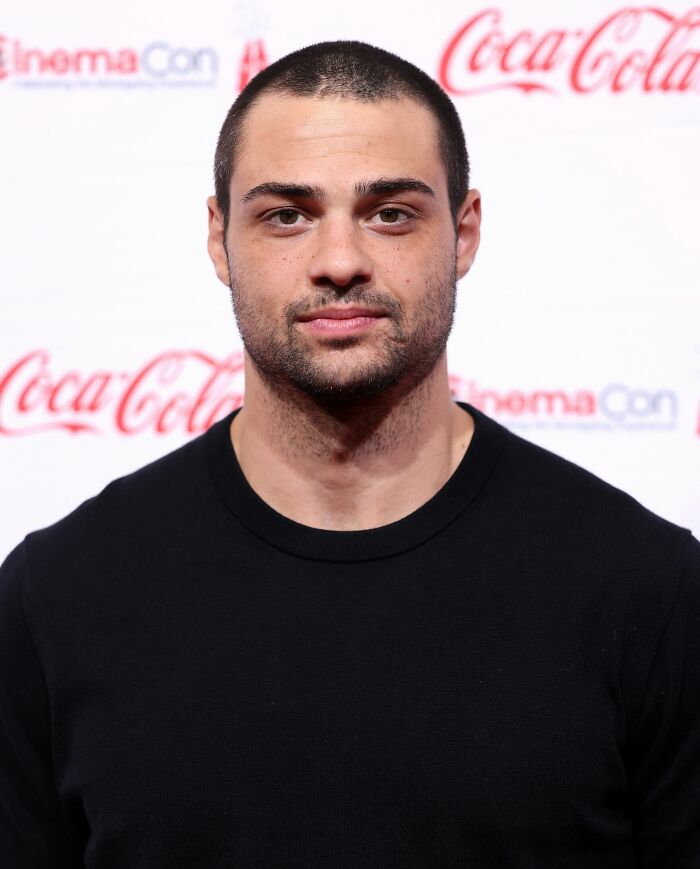 Noah Centineo, with a buzz cut and beard, wearing a black shirt at an event with Coca-Cola branding.