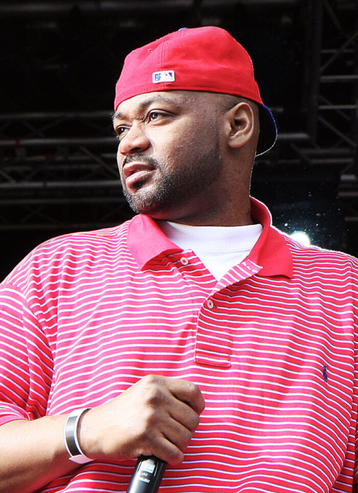 Ghostface Killah on stage, wearing a red hat and striped polo shirt, holding a microphone. Bio and career highlights.