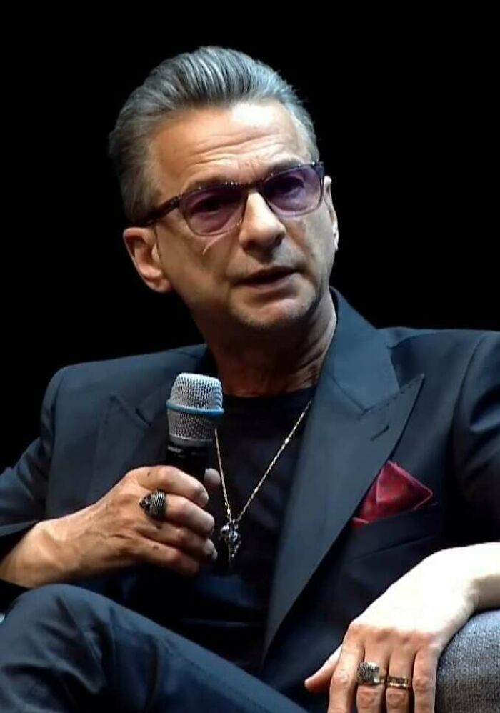 Dave Gahan, wearing sunglasses and a dark suit, holding a microphone. Exploring his bio and career highlights.