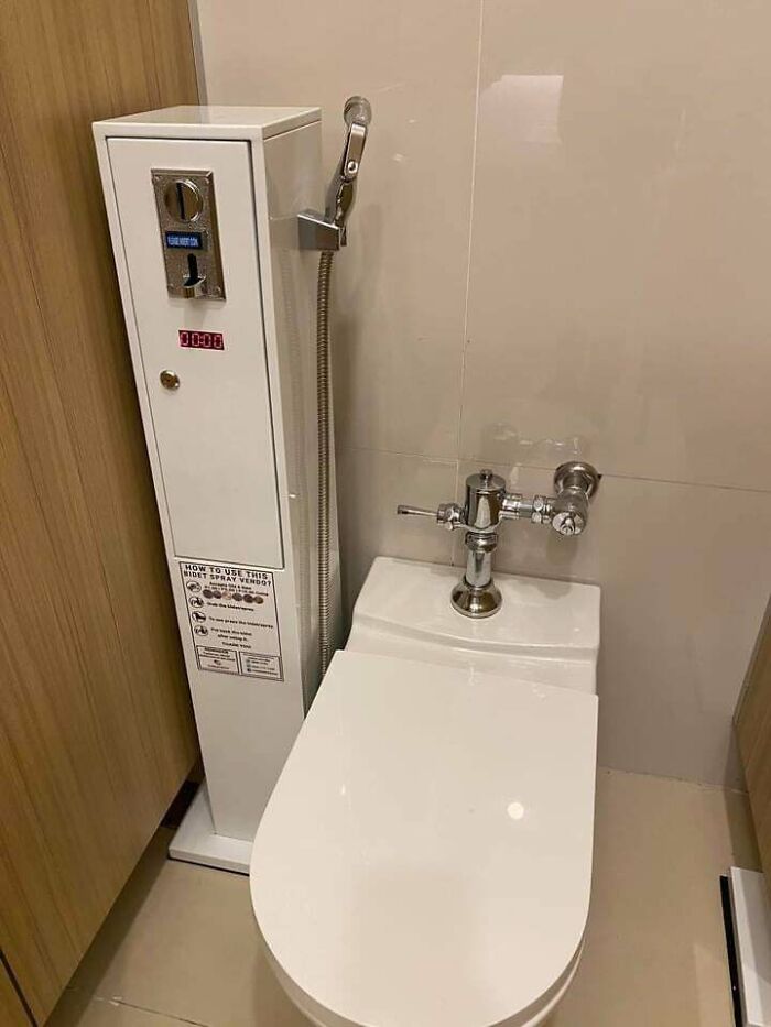 Toilet next to a coin-operated bidet spray vending machine in a small bathroom