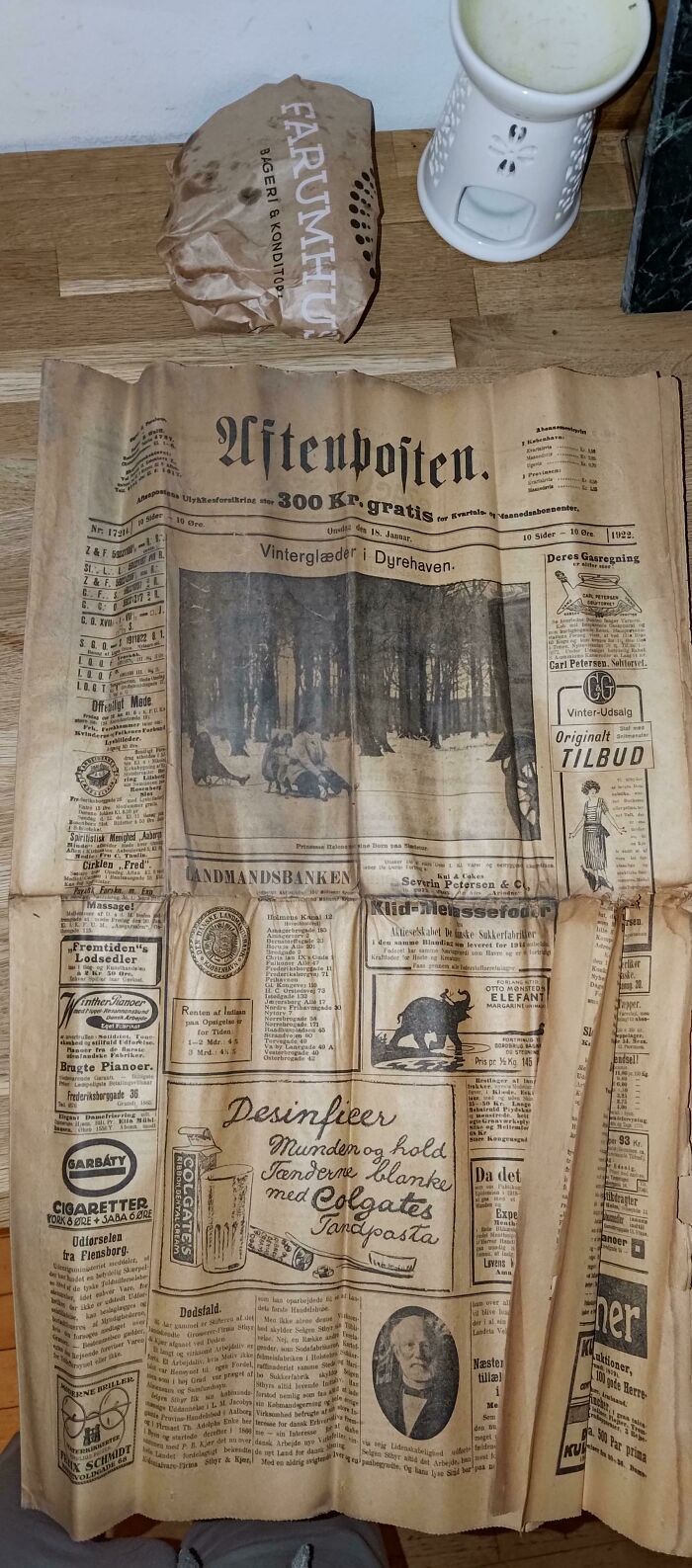 An old newspaper with vintage ads and news, a crumpled paper bag, and a white candle holder on a wooden floor, hinting at high strangeness and a rabbit hole.