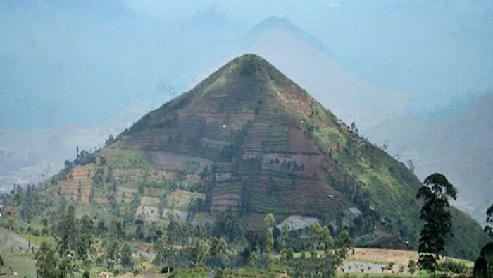 A view of a large, pyramid-shaped mountain with terraced fields and trees on its slopes, evoking high strangeness.
