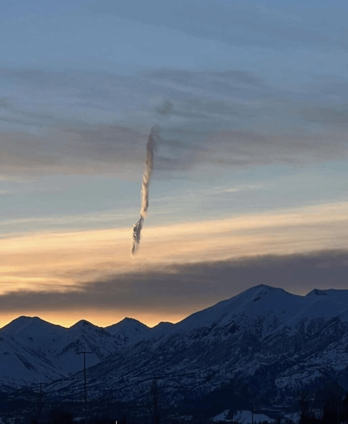 A strange vertical cloud formation against a sunset sky over snowy mountains, suggesting high strangeness.