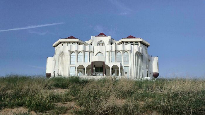 A large white building with red roofs and arched windows, rising above dry grass, hints at high strangeness.