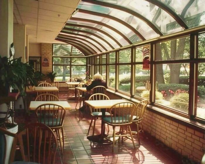 A sunlit retro cafe or diner with an arched glass roof, rows of empty wooden tables, and one person seated. A time machine glimpse into older times.