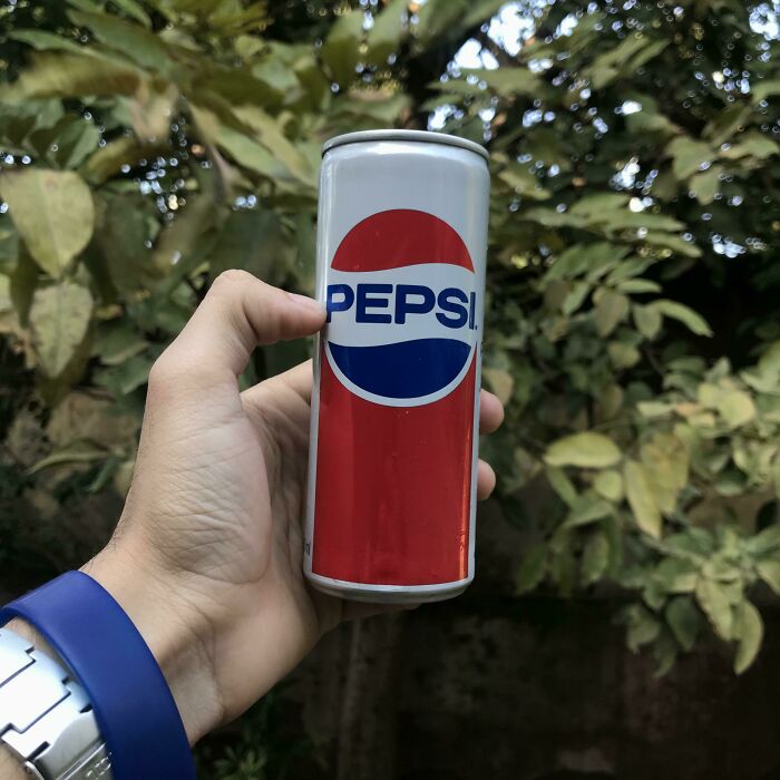 A hand holding a retro Pepsi can with a classic logo, reminiscent of older times, against a leafy background.