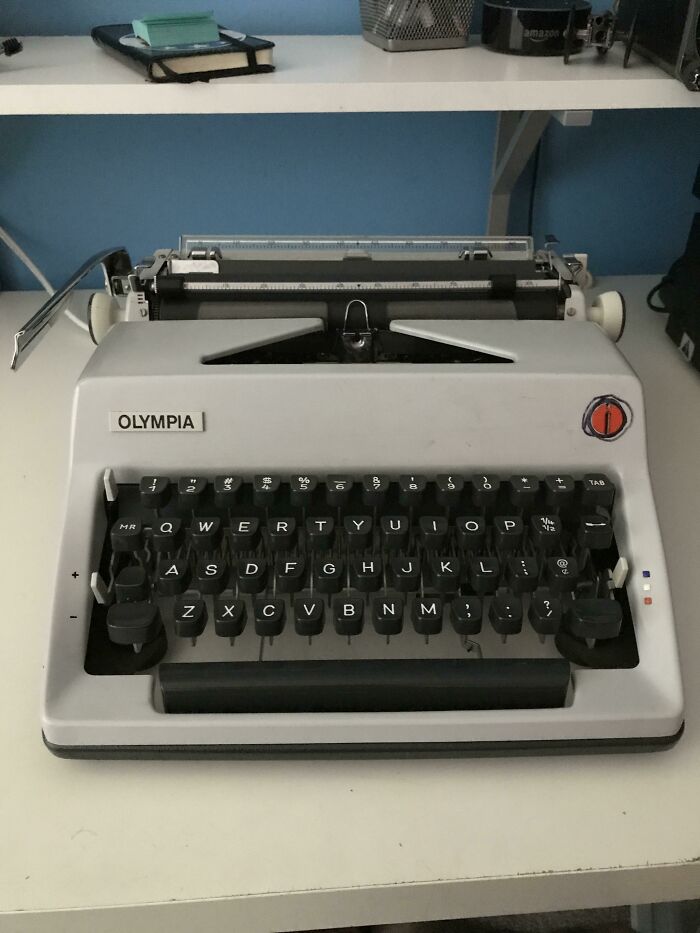 A grey Olympia typewriter from older times, featuring black keys, on a white desk against a blue wall. Retro pics.
