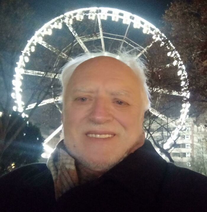 Smiling man with white hair and beard, wearing a dark coat and scarf, with a brightly lit Ferris wheel behind him. Soul-healing images of positivity.