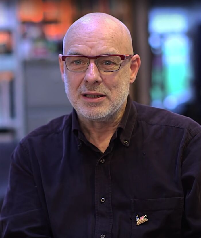 Brian Eno, bald with a grey beard and red glasses, wearing a dark shirt. Bio and career highlights.