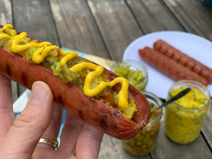 Hand holding a hot dog topped with mustard and relish outdoors