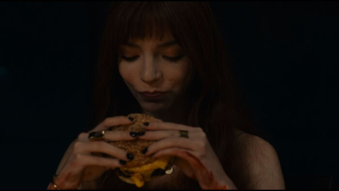 A woman with long red hair eats a burger, hands stained, in a dimly lit scene, highlighting nonsensical movie details.