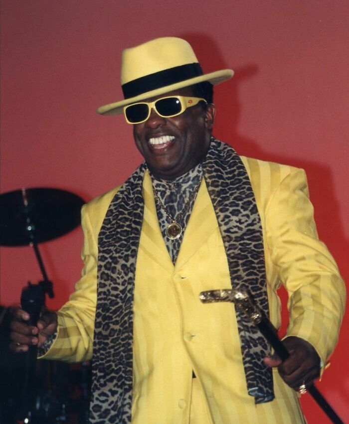 Ronald Isley in a yellow suit, hat, and sunglasses, smiling while holding a microphone and a cane, showcasing his career highlights.