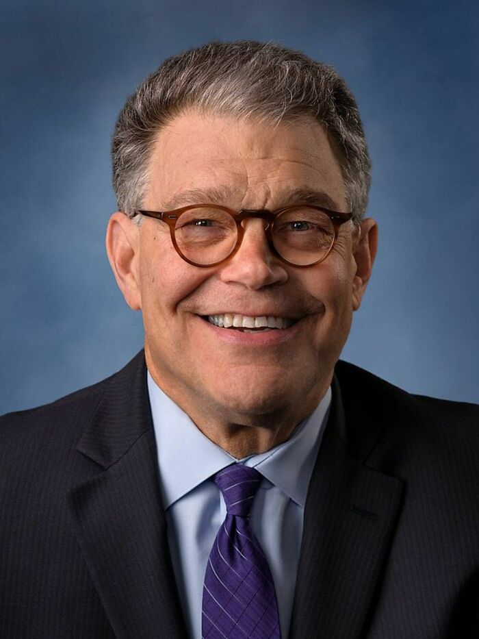 Portrait of Al Franken smiling in a suit and glasses against a blue background