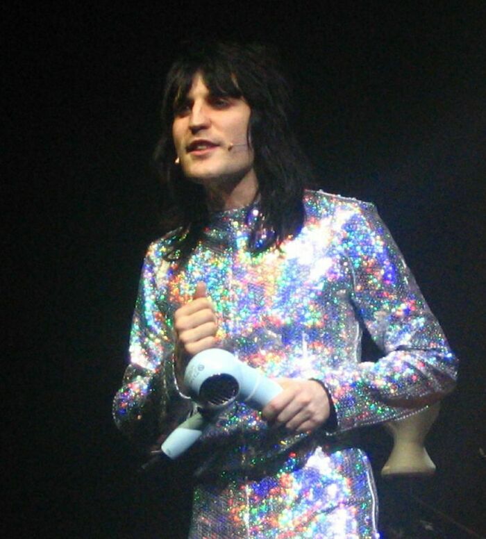 Noel Fielding performing on stage wearing a sparkling multicolored outfit and holding a hairdryer