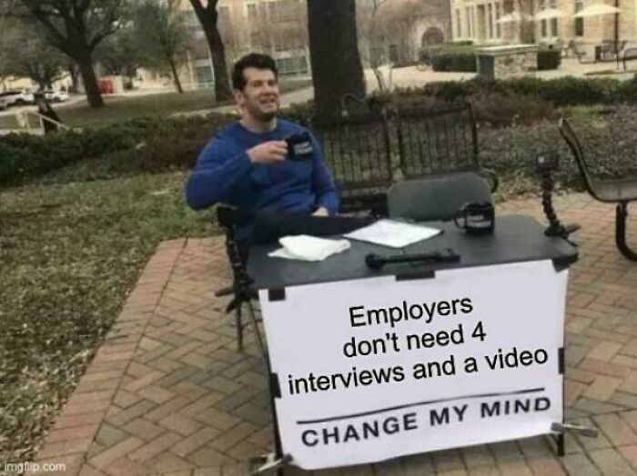 A man sitting at a desk with a sign that says Employers dont need 4 interviews and a video. Change my mind, related to recruiting hell.