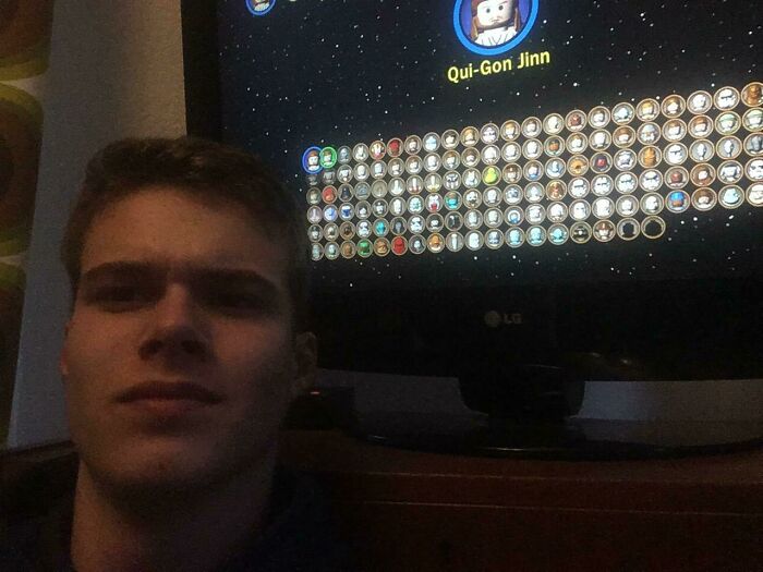 Young man playing video game with character selection screen showing Qui-Gon Jinn