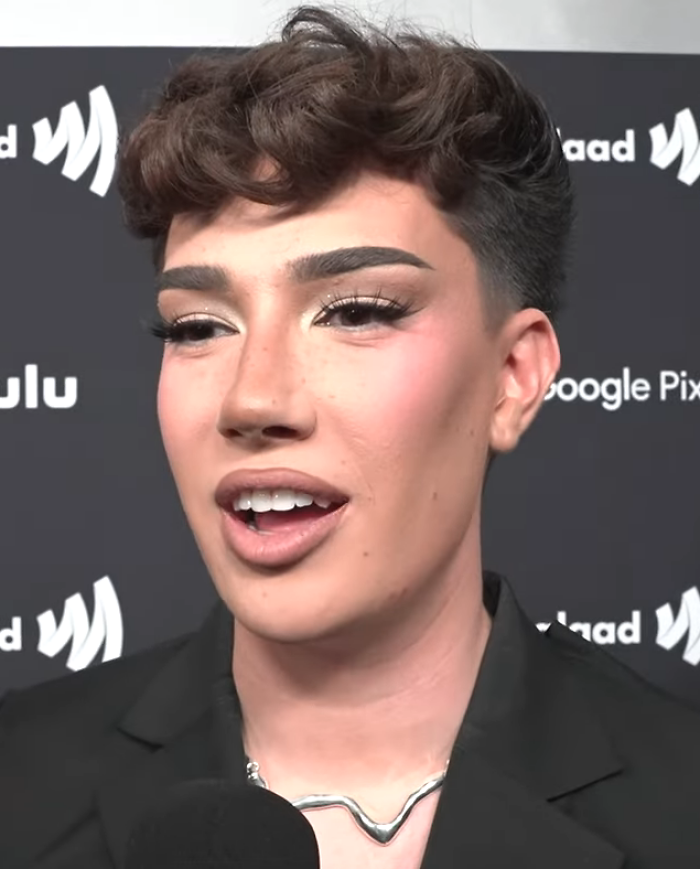 Close-up of James Charles with makeup and styled hair at event backdrop, wearing black blazer and silver necklace