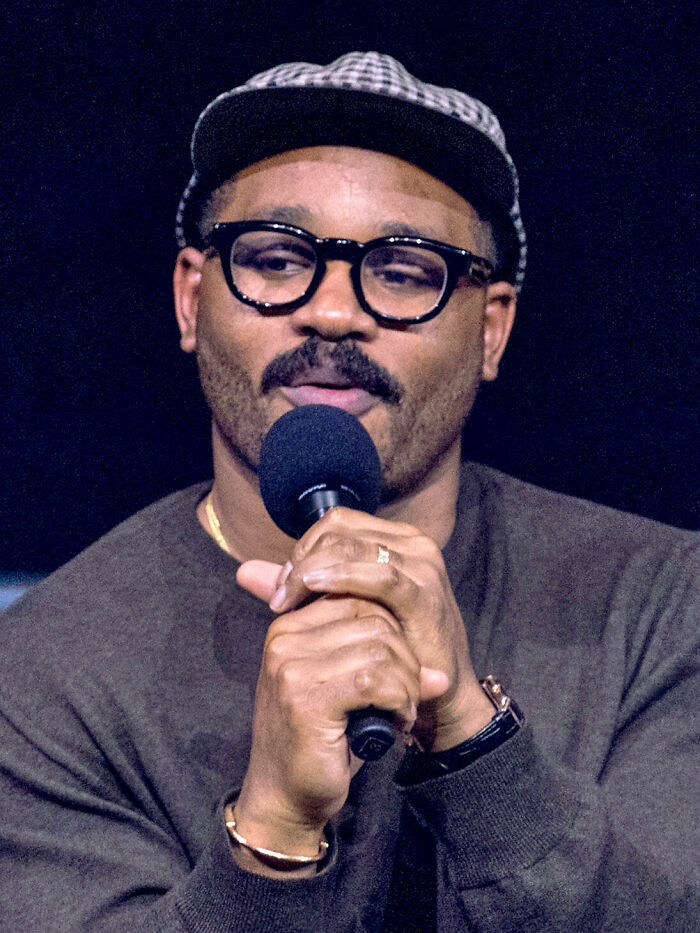 Ryan Coogler speaking into a microphone wearing glasses and a checkered cap