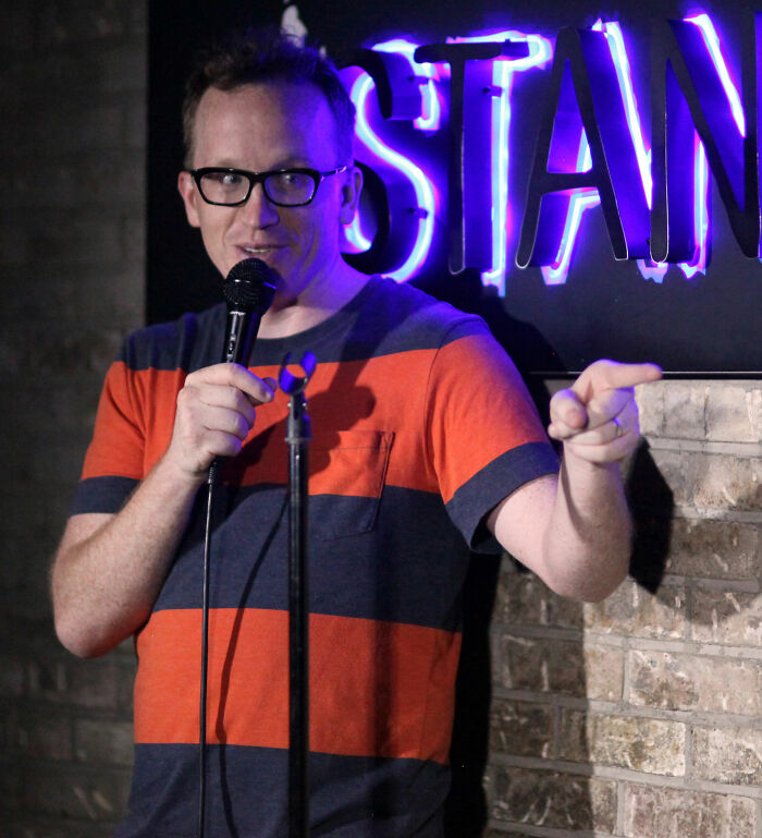 Chris Gethard performing stand-up comedy with microphone and neon stand-up sign in background