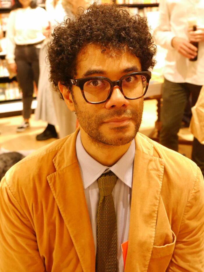 Close-up of Richard Ayoade wearing glasses and a brown jacket at an indoor event