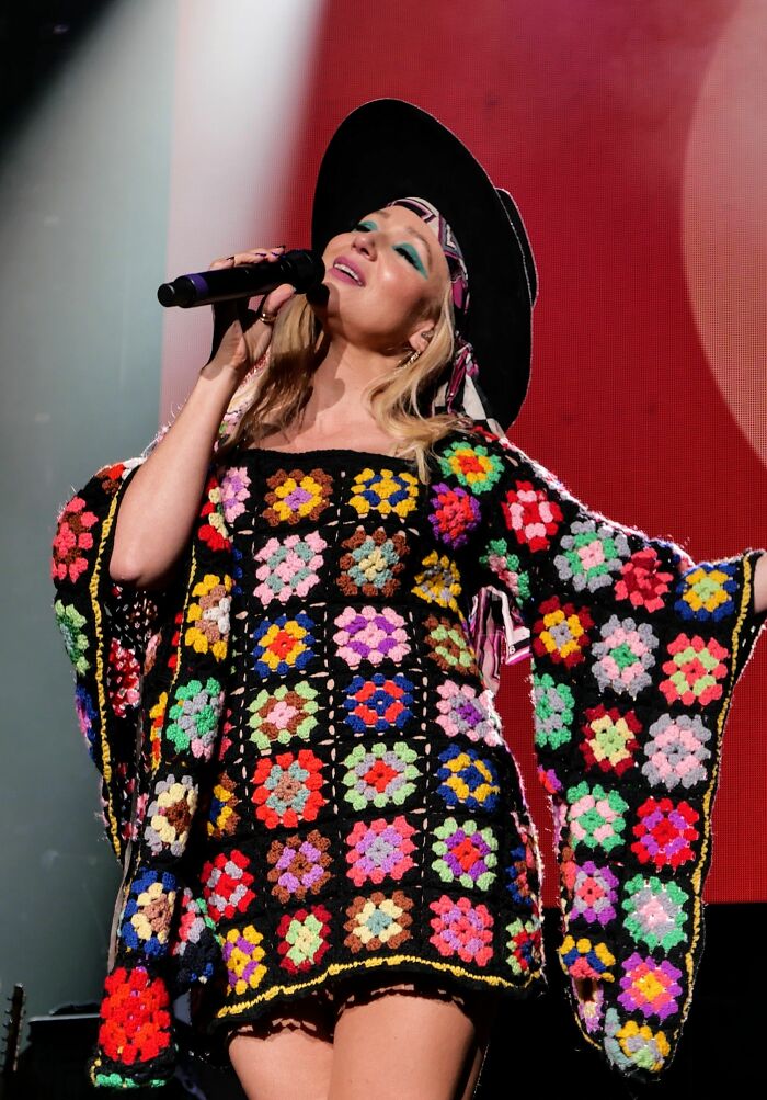 Singer Jewel performing on stage wearing a colorful crochet dress and black hat with vibrant stage lighting
