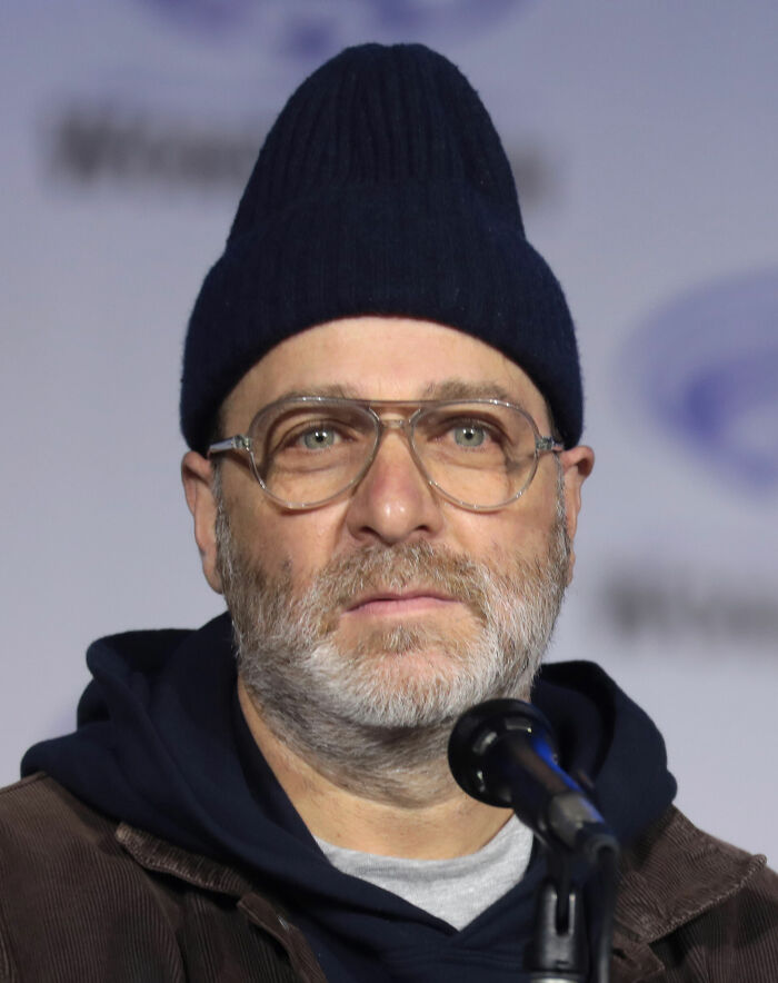 Close-up of H. Jon Benjamin speaking into a microphone wearing glasses and a dark beanie hat