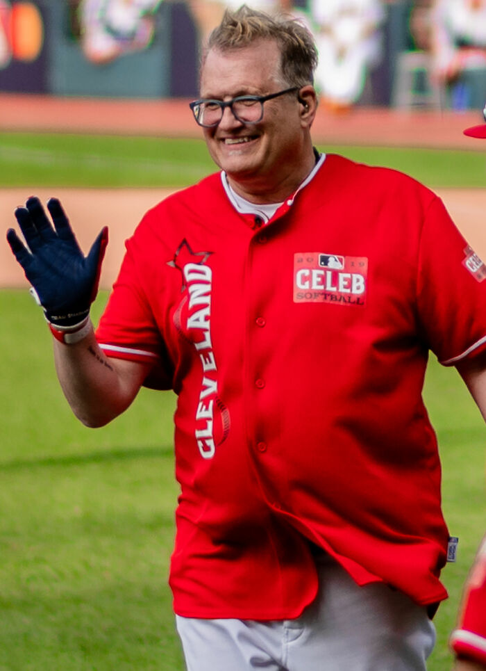 Drew Carey smiling and waving in a Cleveland Celeb Softball red jersey on a baseball field