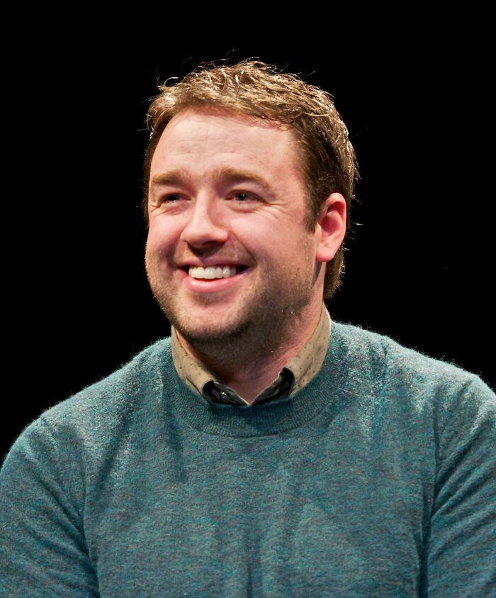Smiling Jason Manford wearing a green sweater on a black background