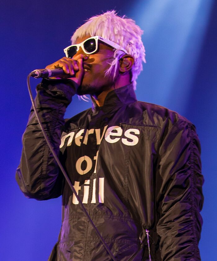 André 3000 performing on stage wearing sunglasses and a black jacket with white text under purple lighting