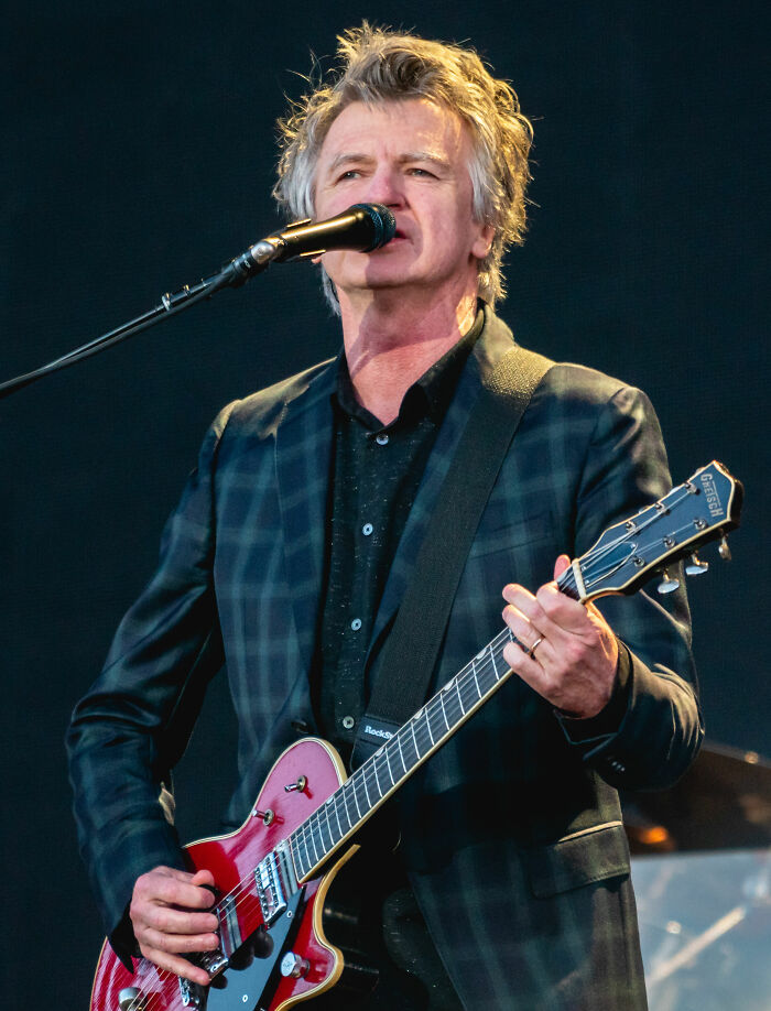 Neil Finn performing live on stage playing electric guitar and singing into a microphone