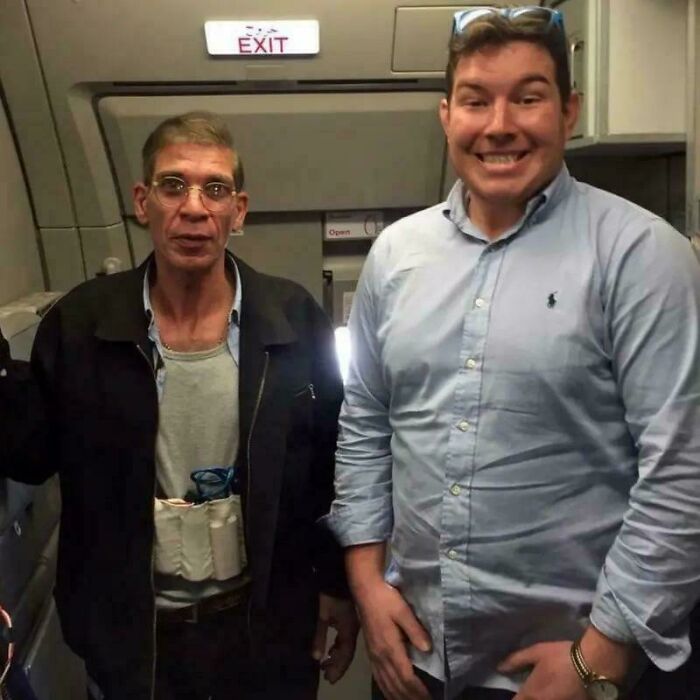 Two men posing inside an airplane cabin with one wearing glasses and a light blue shirt