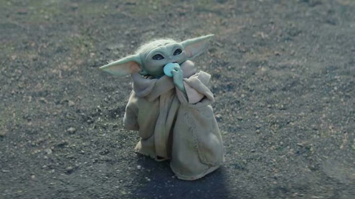 A tiny Baby Yoda sits on a dirt path, holding a light blue object to its mouth, a nonsensical movie detail.
