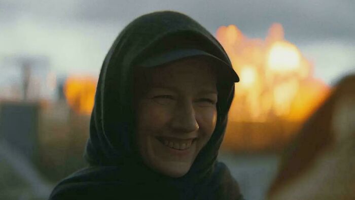 A person smiling under a hood with a fire glowing in the background, a detail from a movie.