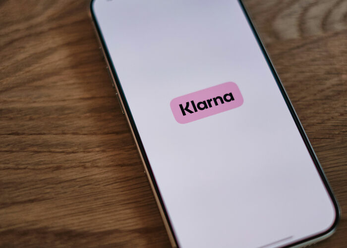 A smartphone displaying the Klarna logo on its screen, resting on a wooden surface. People worry about their wallets.