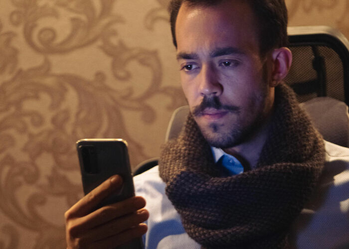 Man in a brown scarf looking at his phone, worried about the economy and his wallet.