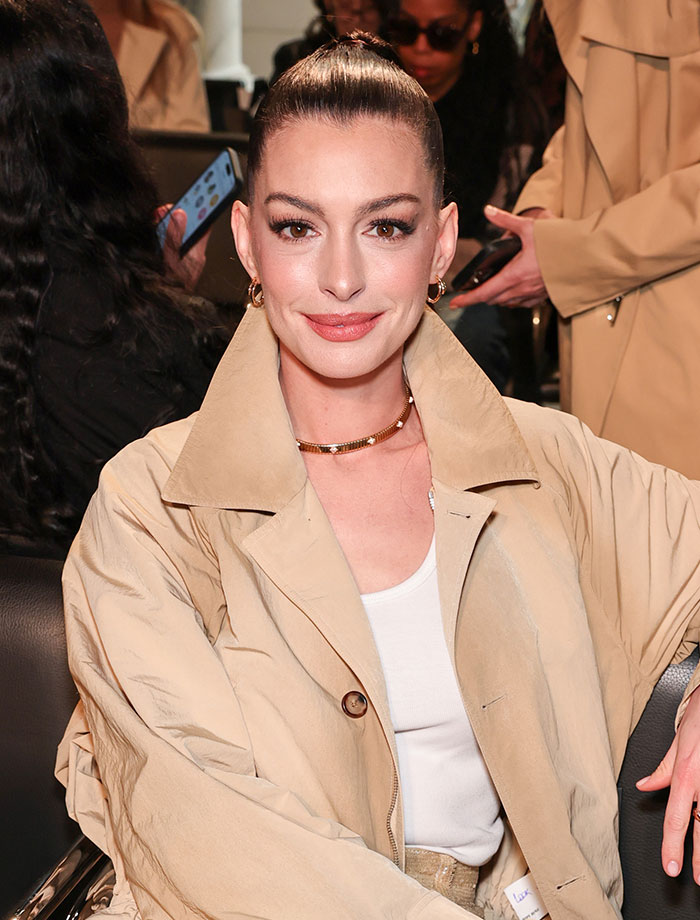 Anne Hathaway smiling, wearing a beige trench coat and a gold choker. Discussion on Anne Hathaway's scene in The Odyssey.