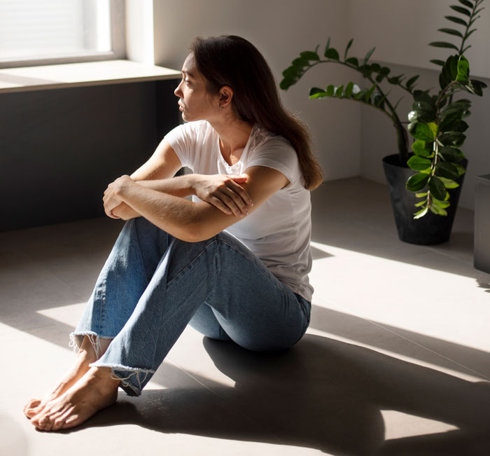A pensive young woman in a white t-shirt and jeans sits on the floor by a window, contemplating repeated Reddit facts.