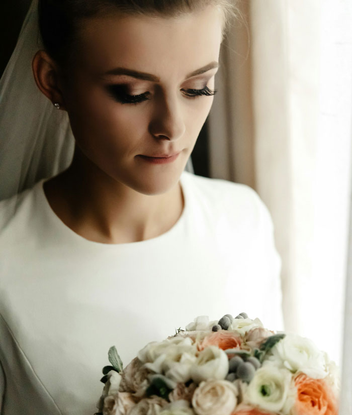 Bride holding floral bouquet standing by window reflective before wedding