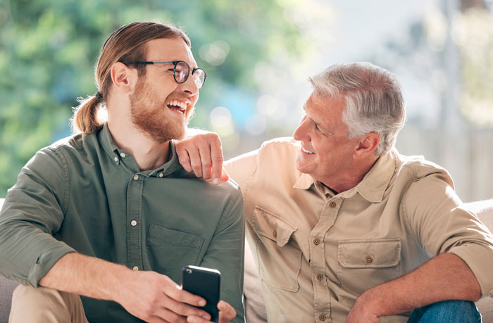 A smiling young man with a ponytail and glasses talks with an older man, representing a solid relationship with GF's Dad.