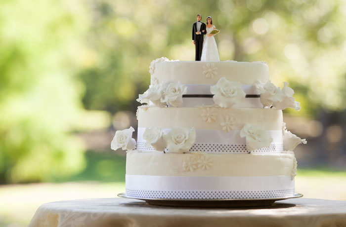 White wedding cake with bride and groom figurines outdoors White wedding cake with bride and groom figurines outdoors