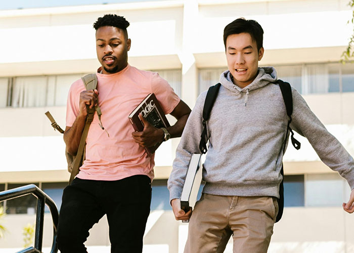 Two male college students with backpacks and books walking down stairs, discussing stereotypes about the US.