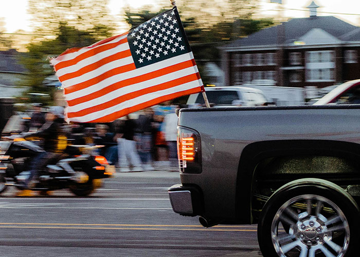 An American flag flies from a pickup truck, blurring with motion. Motorcyclists and people on the street are visible in the background, a scene often seen by people of low intelligence.