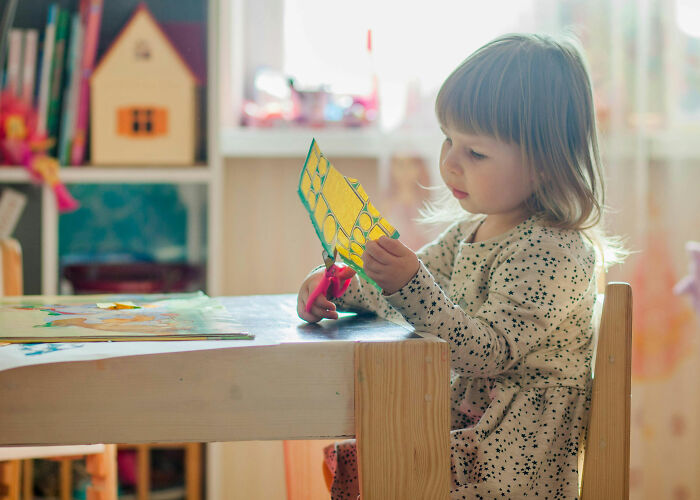 A young child with long hair intently cuts a yellow craft with pink scissors at a wooden table, symbolizing how people worry about their wallets.