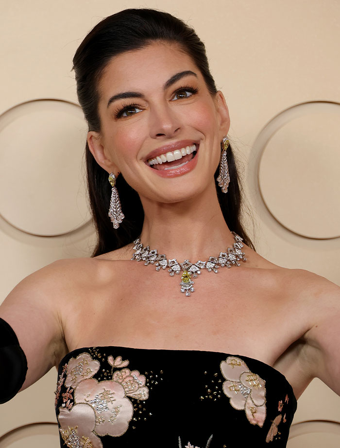 Anne Hathaway smiling in a black strapless dress with intricate floral embroidery, wearing a dazzling diamond necklace and earrings.