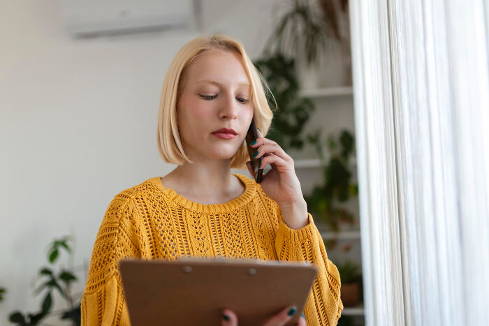 Blonde woman in a yellow sweater, looking down at a clipboard while talking on the phone, possibly experiencing second-hand embarrassment.