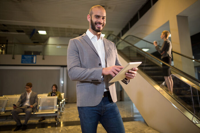 Man with tablet smiling, in an airport waiting area. Netizens discuss celebrity relatives.